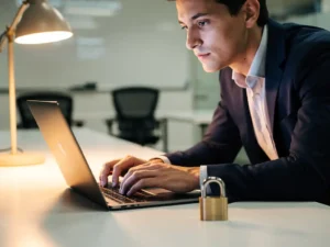 Professional leaning over laptop in modern training room with padlock on desk, warm amber light, navy and gold tones.