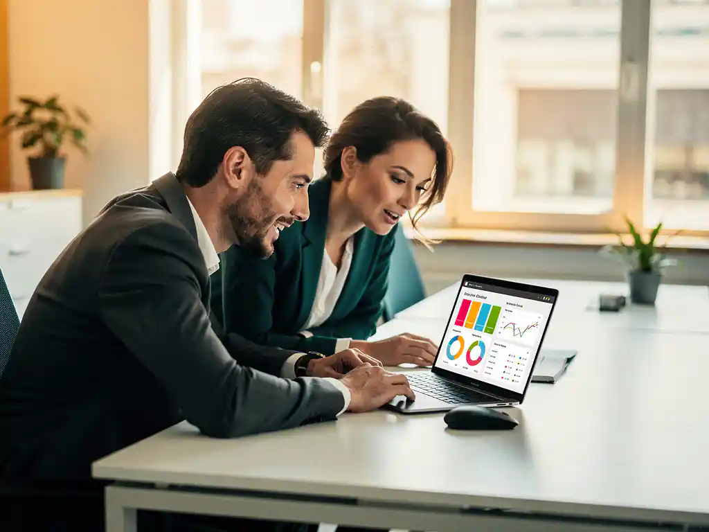 Twee collega's bekijken samen data-dashboards op een laptop aan een modern bureau in een licht kantoor.